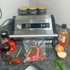 Vacuum sealer on a kitchen counter with raw meat, vegetables, and a bottle of oil.
